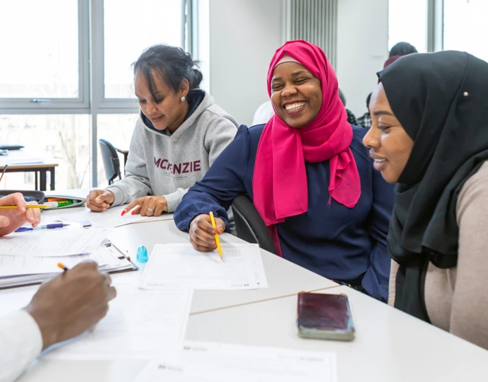 A diverse group of students, including a woman in a pink hijab, laughs and collaborates while working on an assignment in a classroom. A diverse group of students, including a woman in a pink hijab, laughs and collaborates while working on an assignment in a classroom.